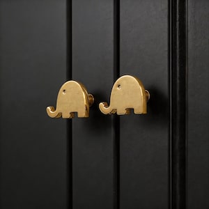 May include: Two gold-coloured elephant-shaped cabinet knobs fixed to a black cabinet door. The knobs have a smooth, polished finish and are designed with a trunk, ears, and feet. The cabinet door has vertical paneling.
