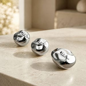 May include: Three polished silver-toned metallic stones with irregular, organic shapes. Each stone features the name Adeline engraved in a black script font, accompanied by a small heart symbol above the letter i, creating a reflective surface.