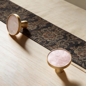 Op de afbeelding: Twee kastknoppen met een gouden afwerking en een ronde, lichtroze, gemarmerde kern. De knoppen staan op een licht houten oppervlak met een donkere, bedrukte loper. De knoppen zijn waarschijnlijk voor meubels.