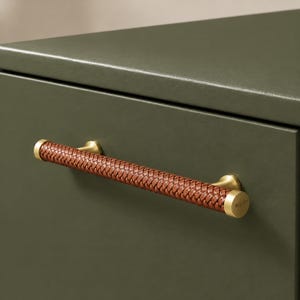 Op de afbeelding: Een ladehandgreep met een cilindrische stang omwikkeld met bruin gevlochten leer, bevestigd door twee eindkappen en steunen van geborsteld messing op een effen mat olijfgroen ladefront van een kast.