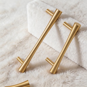 May include: Gold-colored cabinet pulls with a modern design. The pulls have a cylindrical bar with conical ends and a perpendicular handle. The hardware is displayed on a white, fluffy surface.