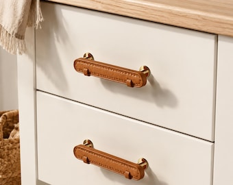 Può includere: Due maniglie per cassetti orizzontali con un rivestimento in similpelle color cuoio, fissate con finiture in metallo dorato. Le maniglie sono montate su un mobile bianco con una superficie in legno chiaro visibile in alto.