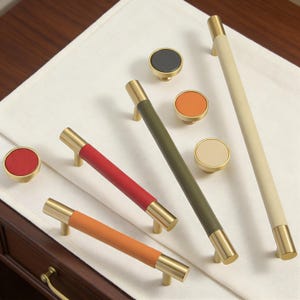 May include: A collection of cabinet hardware, including gold-toned handles and knobs. The handles feature colored leather wraps in red, orange, and olive green. Knobs have matching leather in red, orange, cream, and black. The hardware is displayed on a white cloth.