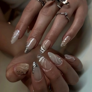 May include: Close-up of a hand with long, almond-shaped nails. The nails feature a variety of designs, including silver metallic accents, swirling patterns, and clear gel with embedded gems. Rings adorn the fingers, including a bow-shaped ring.