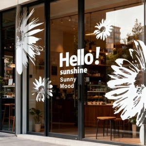 Peut inclure: Devanture de magasin avec portes et fenêtres en verre ornées de motifs de marguerites blanches et des mots "Hello! sunshine Sunny Mood". L'intérieur est visible, avec des meubles et des étagères en bois.