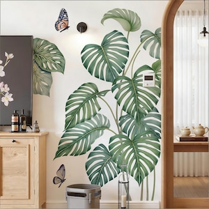 Op de afbeelding: Een muursticker met grote, groene monstera bladeren met witte aderen. Er zijn ook twee vlinderstickers aanwezig. De bladeren zijn zo gerangschikt dat ze een tropisch plantontwerp creëren.