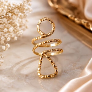 May include: A gold-toned ring featuring a textured, spiraling design that transitions from a circular top loop into a triangular base. The metal surface has a hammered or dimpled finish, creating a reflective appearance throughout the piece.
