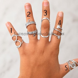 May include: A hand showcasing five silver rings. Ring 1 has two connected circles, ring 2 features a spiral design, ring 3 is a long oval, ring 4 has heart accents, and ring 5 has a spiral design. The rings are of different styles.