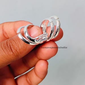 Può includere: Un anello color argento dal design unico, con più fasce collegate. L'anello è tenuto tra le dita su uno sfondo grigio chiaro. L'anello ha una superficie lucida e riflettente.