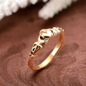 May include: A gold Claddagh ring. The ring features a heart held by two hands, with a crown above the heart. The ring is set against a brown background.