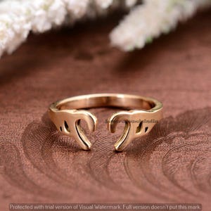 Puede incluir: Un anillo dorado con un diseño abierto. El anillo presenta dos formas curvas estilizadas que se encuentran en el centro. El anillo está sobre un fondo marrón, con una marca de agua en la esquina inferior derecha.