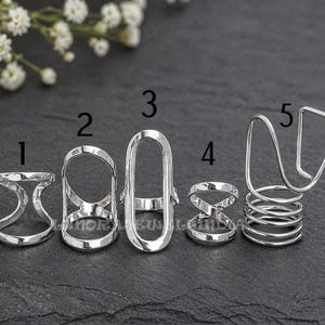 Op de afbeelding: Vijf zilverkleurige ringen in verschillende ontwerpen worden getoond. De ontwerpen omvatten een open X, een ovaal en een spiraal. De ringen zijn waarschijnlijk van metaal en geschikt voor dagelijks gebruik. Ze worden op een donkere ondergrond gepresenteerd.