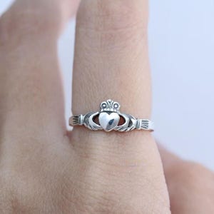 May include: A silver Claddagh ring with a heart, crown, and hands design. The heart is the central element, flanked by hands and topped with a crown. The ring is worn on a finger.