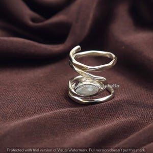 May include: A silver-colored ring with a spiral design. The ring features a central, light-colored stone and a textured band. The ring is displayed on a brown fabric background.