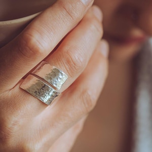 Puede incluir: Un anillo abierto de color plateado con una banda ancha y un acabado texturizado martillado. El anillo tiene dos extremos distintos ligeramente curvados que no se unen, creando un diseño ajustable que luce un brillo metálico característico.