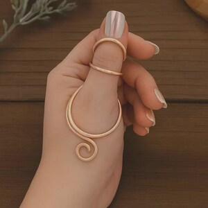 May include: A hand wearing three gold rings. One ring is on the index finger, another is on the middle finger, and the third ring is a large, curved design that wraps around the middle finger and extends down the hand.