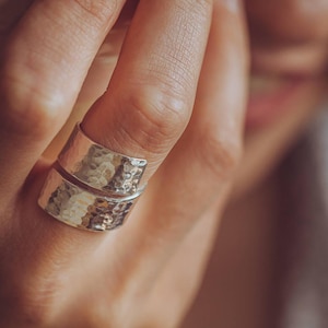 Puede incluir: Un anillo ancho de plata que presenta una textura martillada distintiva en toda su superficie. La banda tiene un diseño envolvente que crea la apariencia de dos anillos apilados, con un acabado metálico pulido que refleja la luz.