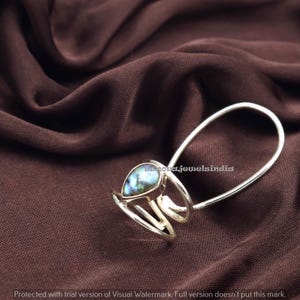 Peut inclure: Une bague en argent ornée d'une pierre précieuse en forme de poire aux couleurs irisées. La bague présente un design unique avec une boucle et deux bandes. La pierre affiche des nuances de bleu et de vert. Le fond est un tissu marron foncé.
