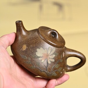 May include: A small, brown ceramic teapot with a lid and handle. The teapot is decorated with a white flower and green leaves. The teapot is held in a hand.