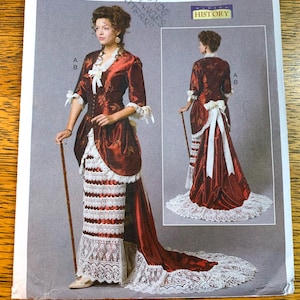 May include: A vintage sewing pattern for a historical costume. The dress is a rich burgundy with white lace and trim. The pattern shows a full-length gown with a long train and a matching jacket. There is also a smaller inset image of the dress on the pattern.