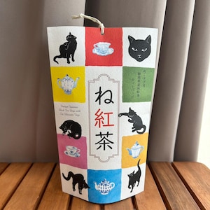 Puede incluir: Un paquete decorativo de bolsitas de té con un diseño de cuadrícula, con siluetas de gatos negros, tazas de té y teteras en cuadrados de colores. El paquete tiene texto japonés.