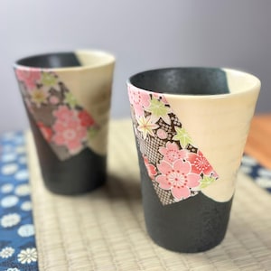 May include: Two ceramic tumblers with a black base, cream-colored upper section, and a diagonal floral pattern featuring pink cherry blossoms. The tumblers have a slightly tapered shape and are set against a neutral background.