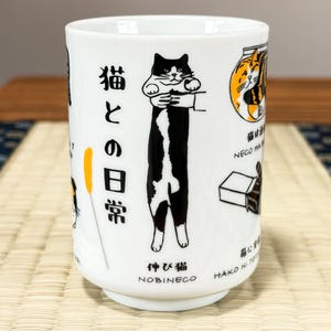May include: A white ceramic mug featuring black and orange illustrations of cats in various poses. The text includes Japanese characters and the English words NOBINECO. One graphic shows a long black and white cat being held vertically.