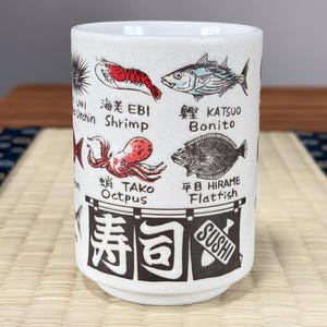 May include: A white ceramic cup with a speckled finish featuring illustrations of various seafood including shrimp, bonito, and octopus. The cup displays Japanese kanji and English text labels for each item, with a large banner at the bottom reading Sushi.