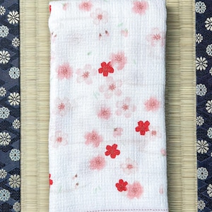 以下が含まれることがあります： ピンクと赤の桜の花柄の白い折り畳みタオル。タオルは、白い花の模様が施された濃い青色の縁取りのある、明るいベージュ色のマットの上に置かれています。背景は木製の表面です。
