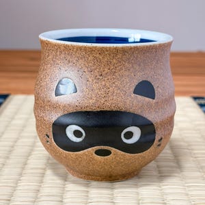 Puede incluir: Una taza de cerámica redondeada con un exterior beige moteado y un interior azul profundo. El frente presenta un diseño de máscara negra con ojos blancos, una pequeña nariz negra y dos orejas negras en la parte superior, similar a un tanuki.