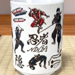 May include: A white ceramic cup with a speckled finish featuring illustrations of ninjas in various poses, scrolls, and weapons. The design includes the Japanese kanji for ninja and the English word NINJA printed in black ink on the front surface.