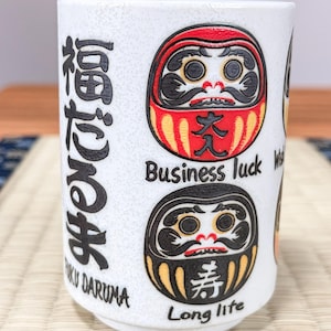 Peut inclure: Un mug en céramique blanche orné de deux illustrations de poupées Daruma. La poupée supérieure est rouge avec le texte Business luck, et celle du bas est noire avec le texte Long life. Des caractères japonais et FUKU DARUMA sont inscrits.