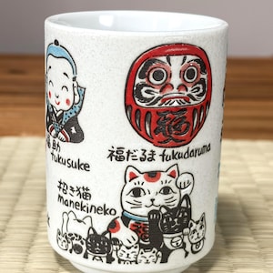 以下が含まれることがあります： 斑点模様の白い陶器製カップ。福助人形、赤い達磨、招き猫のイラストが描かれています。それぞれのイラストの横には、黒い文字で「福助」「福だるま」「招き猫」およびローマ字表記のfukusuke、fukudaruma、manekinekoがプリントされています。