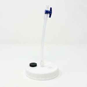 May include: A white circular lid featuring a clear plastic tube extending vertically from the center. The tube is topped with a small blue plastic valve handle. A solid black circular cap is positioned on the surface of the white lid.