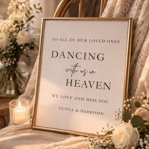 May include: A framed sign with gold trim, displaying the text "DANCING with us in HEAVEN". The sign is propped on a chair draped with a cream-colored blanket, with flowers and a lit candle nearby. The text also reads "TO ALL OF OUR LOVED ONES" and "WE LOVE AND MISS YOU, OLIVIA & HARRISON".