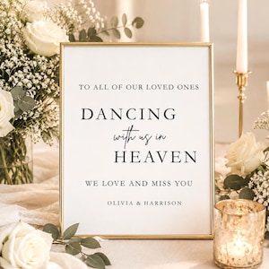 May include: A framed sign with gold trim and the text "TO ALL OF OUR LOVED ONES, DANCING with us in HEAVEN, WE LOVE AND MISS YOU, OLIVIA & HARRISON." The sign is surrounded by white flowers, candles, and a candle holder.