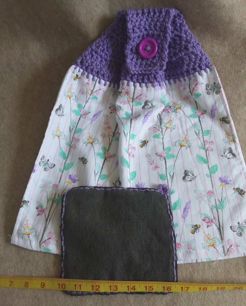 May include: Purple crocheted dish towel with a floral print and a purple button. The towel has a gray fabric insert.