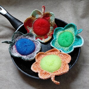 May include: Four colorful, handmade crochet pot scrubbers in a frying pan. Each scrubber is flower-shaped with a central sponge. Colors include red, blue, green, and orange, with coordinating crochet edging. The pan has a black handle.