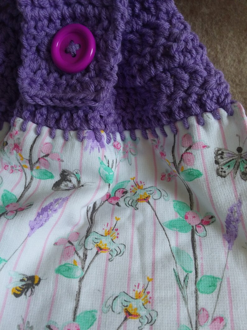 May include: Purple crocheted dishcloth with a pink button and a white fabric backing with a floral pattern.