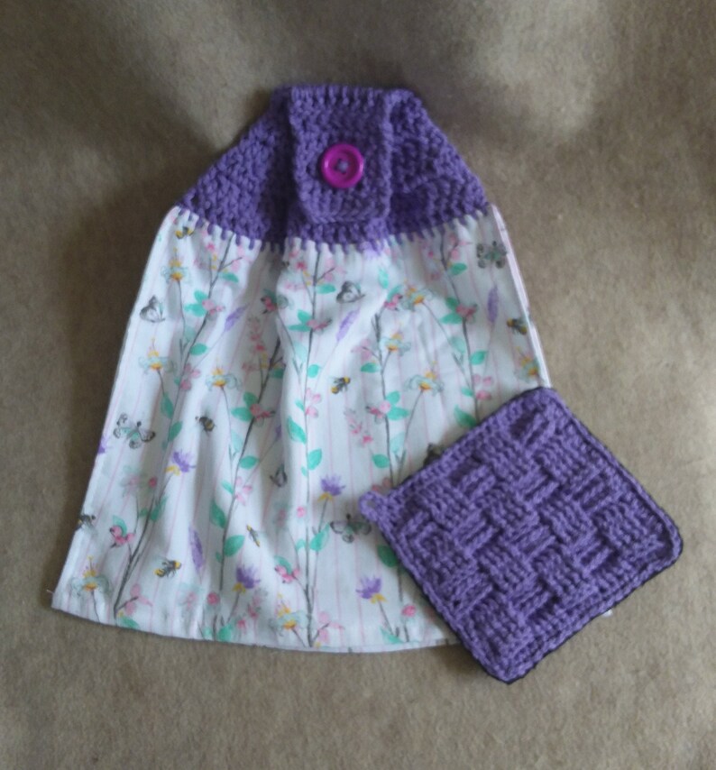 May include: Purple crocheted kitchen towel with a floral print and a matching purple crocheted dishcloth. The towel has a button on the top.
