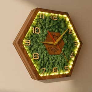 Könnte beinhalten: Sechseckige Wanduhr mit Holzrahmen und einem grünen, moosartigen Innenraum. Die Uhr hat goldfarbene Zeiger und Ziffern mit dem Text "Live For You" auf dem Zifferblatt. Die Uhr wird von warmweißen Lichtern um das Moos herum beleuchtet.