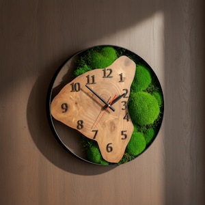 May include: A circular wall clock featuring a wooden face with black numerals and black and red hands. The clock is set within a black metal frame, surrounded by green moss. The clock is mounted on a wooden wall.