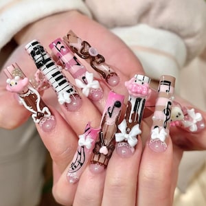 Pode incluir: Um conjunto de unhas compridas e decoradas. Os desenhos incluem um teclado de piano, notas musicais, laços e personagens de desenhos animados. As cores incluem rosa, castanho, branco e preto. As unhas têm um acabamento brilhante.