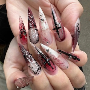 Könnte beinhalten: Eine Hand, die lange, spitze Acrylnägel zeigt. Die Nägel weisen eine Vielzahl von Designs auf, darunter schwarze Filigranarbeiten, Texte mit der Aufschrift "Paradise" und "Heavenly" sowie Miniatur-Schwert- und Kreuzverzierungen. Die Farben sind Rot, Weiß und Transparent.