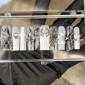 Puede incluir: Un conjunto de diez uñas artificiales en una caja de plástico transparente. Las uñas son principalmente blancas con detalles en negro y adornos de metal plateado, incluyendo anillos, púas y tachuelas. El diseño es atrevido y moderno.