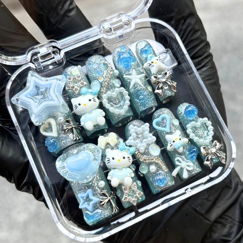 Op de afbeelding: Een set kunstnagels in een doorzichtige etui. De nagels zijn versierd met glitter, blauwe en zilveren accenten, ster- en hartvormen, strikken en kleine Hello Kitty-figuren. De nagels zijn in verschillende tinten blauw.
