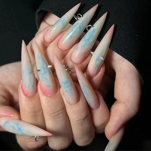 May include: Long, pointed acrylic nails with a gradient design. The nails transition from a pink base to a white tip, with blue cloud-like patterns. Some nails have silver rings and studs.
