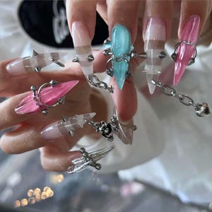 May include: Long, pointed artificial nails featuring a translucent finish in shades of pink, blue, and clear. Each nail is adorned with silver-toned metal hardware, including small spikes, circular rings, and a delicate chain with a heart charm.