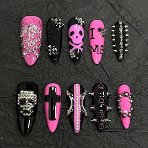 May include: A set of ten press-on nails featuring a black and pink color palette. Designs include a pink marble nail with rhinestones, black nails with silver studs, a pink skull design, a pink nail with the text I ME, and a black cross design.