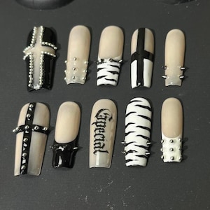May include: A set of ten press-on nails featuring a nude base with black and white designs. Details include silver studs, a black cross, zebra stripes, and the word Special in black gothic lettering. The nails have a long, rectangular shape.